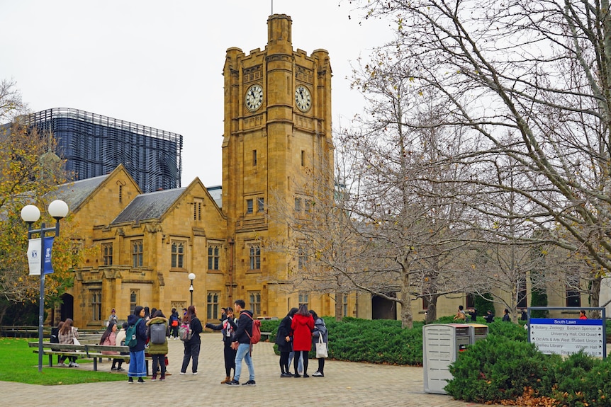 About a dozen student are seen talking in a courtyard, in front of a tall sandstone clock tower at the University of Melbourne.