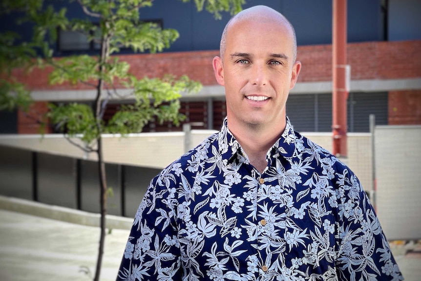 Man with shaved head and Hawaiian shirt stands in front of building and tree.