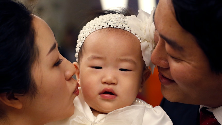 A baby wearing a white headband receives kisses on the cheek from her mother and father on each side. 