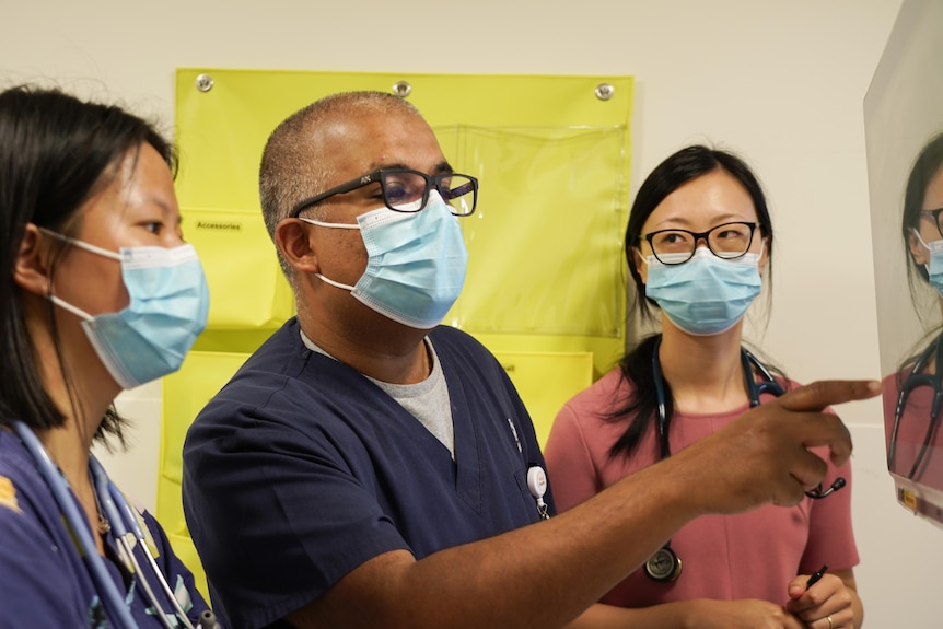 A male doctor wearing a mask points at a screen while two female doctors watch on